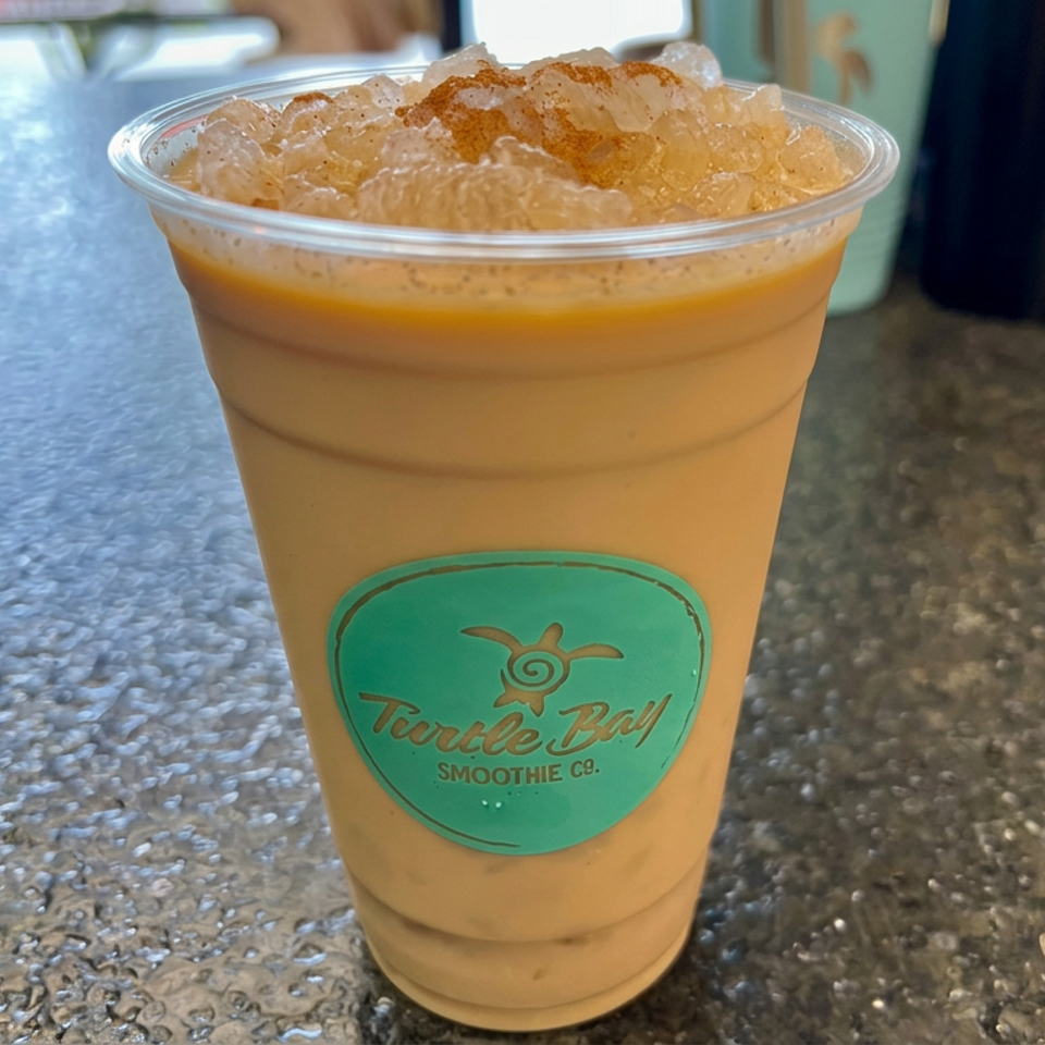 Clear cup of iced caramel-colored smoothie topped with crushed ice, featuring a Turtle Bay Smoothie Co. logo on a café counter.
